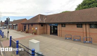 A single-storey building in red brick with a low sloping roof. There is a paved area in front of the building with blue bicycle racks and a blue and black fence.
