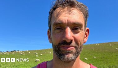Nature Friendly Farming Network member Rhodri Lloyd-Williams stands in a field of sheep on his farm near Talybont, Ceredigion.  He has short, dark hair and a beard and is wearing a pink vest.  He is seen standing against a backdrop of a green field and blue sky.