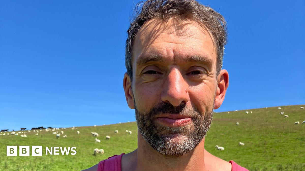 Nature Friendly Farming Network member Rhodri Lloyd-Williams stands in a field of sheep on his farm near Talybont, Ceredigion.  He has short, dark hair and a beard and is wearing a pink vest.  He is seen standing against a backdrop of a green field and blue sky.