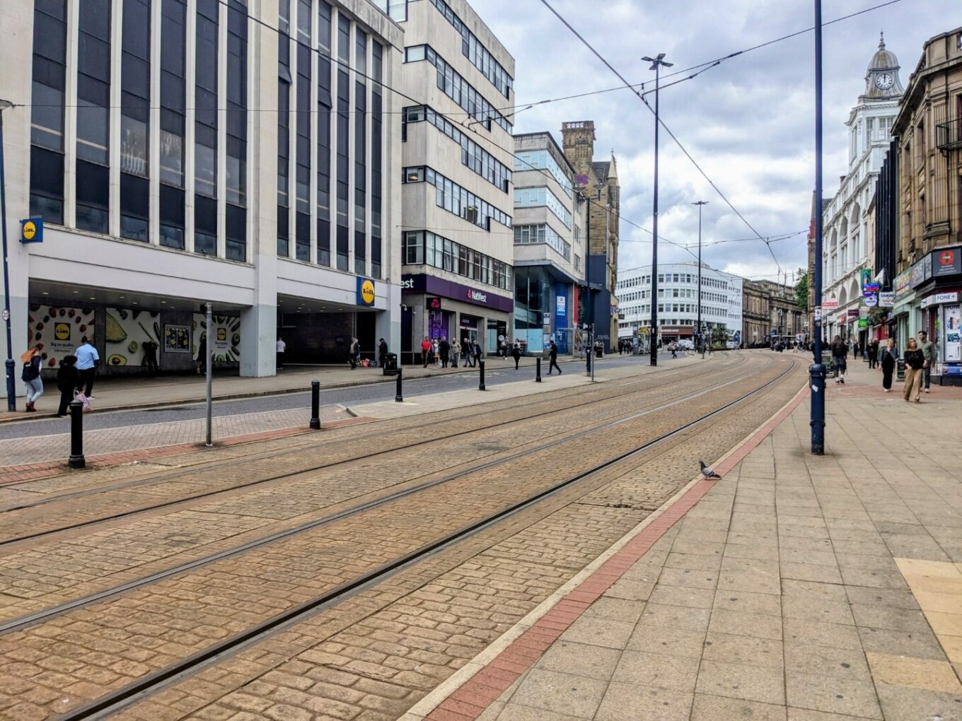 A photo of a city centre street in the middle of the day, with tall buildings on each side and two tram lines running down the middle