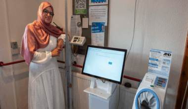 Obaida is standing inside a surgery next to one of the new machines. She is wearing a hijab and a white dress and glasses. She is leaning on equipment next to the machine which is white with a computer screen above it.