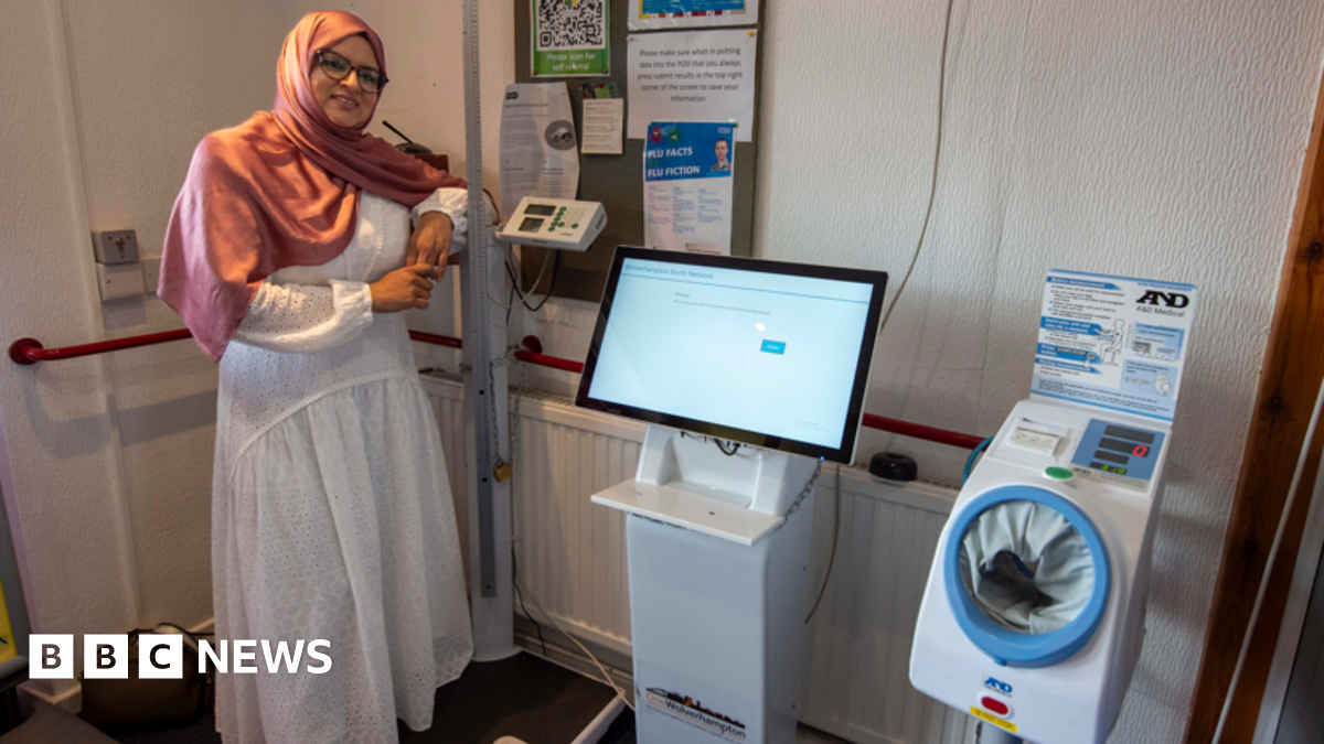 Obaida is standing inside a surgery next to one of the new machines. She is wearing a hijab and a white dress and glasses. She is leaning on equipment next to the machine which is white with a computer screen above it.