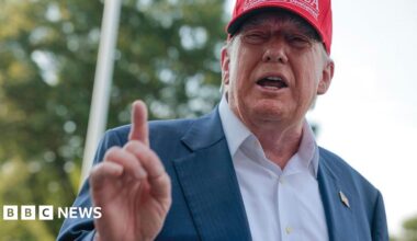 Donald Trump wearing a white shirt and navy suit jacket with no tie. He is wearing a red cap which bears the words: GULF OF AMERICA. Yet another TRUMP development.