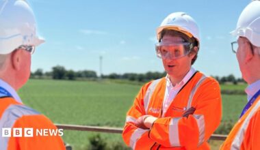 Steffan Aquarone pictured facing towards the camera. He is wearing an orange fluorescent jacket, and PPE including gloves, hard hat and goggles. He is standing outside on a balcony talking to two people from Anglian Water. Both men he is talking to are also dressed in orange fluorescent jackets with the Anglian Water logo on the back, hard hats and goggles.