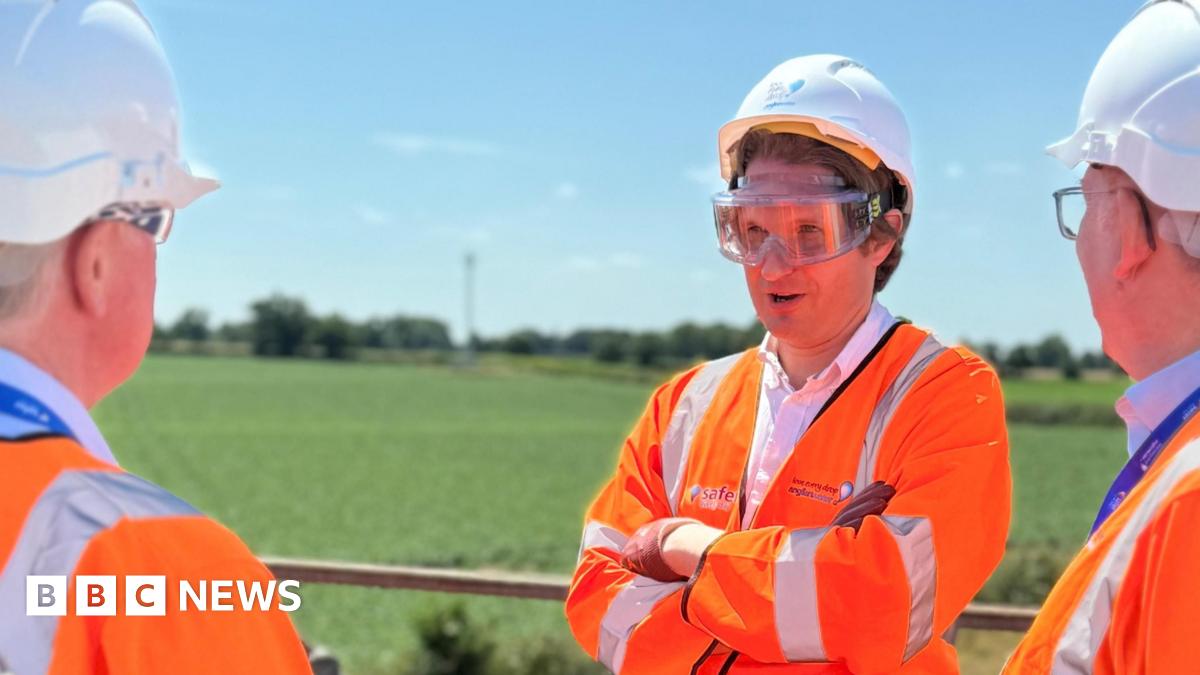 Steffan Aquarone pictured facing towards the camera. He is wearing an orange fluorescent jacket, and PPE including gloves, hard hat and goggles. He is standing outside on a balcony talking to two people from Anglian Water. Both men he is talking to are also dressed in orange fluorescent jackets with the Anglian Water logo on the back, hard hats and goggles.