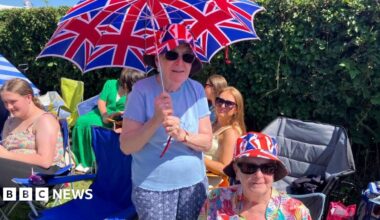 Two ladies are at the side of the road. One is sitting and wearing a Union flag bucket hat and a floral shirt. The other is standing over her holding a Union flag umbrella. She is wearing the same hat and a blue top and trousers.