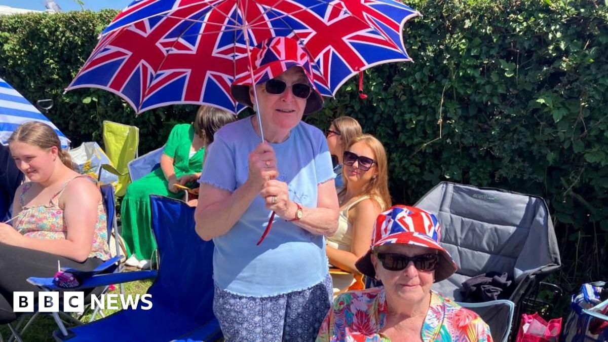 Two ladies are at the side of the road. One is sitting and wearing a Union flag bucket hat and a floral shirt. The other is standing over her holding a Union flag umbrella. She is wearing the same hat and a blue top and trousers.