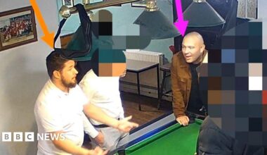 A screenshot from CCTV footage showing a group of men gathered around a pool table in a pub. The man on the left, with dark hair and wearing a white t-shirt, has been highlighted with an orange arrow pointing at his head. The second man on the right, who is bald and wearing a black t-shirt and brown jacket, is highlighted with a pink arrow. The other people in the image have been intentionally blurred out.