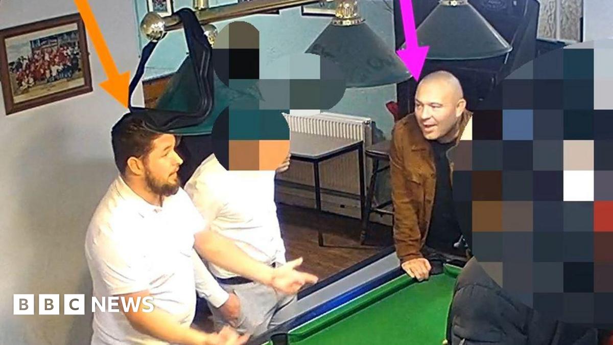A screenshot from CCTV footage showing a group of men gathered around a pool table in a pub. The man on the left, with dark hair and wearing a white t-shirt, has been highlighted with an orange arrow pointing at his head. The second man on the right, who is bald and wearing a black t-shirt and brown jacket, is highlighted with a pink arrow. The other people in the image have been intentionally blurred out.