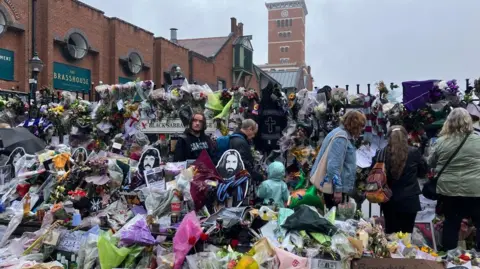 Flowers and notes fill the majority of the image, with some reaching the top of tall black railings. Five people are by the tributes. Buildings are in the background at the top of the photo. 