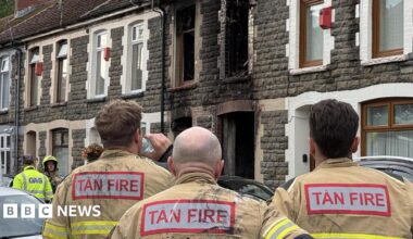 A stone terraced house with fire damage around the windows and doors. Three fire officers look at the house with their backs to the camera.