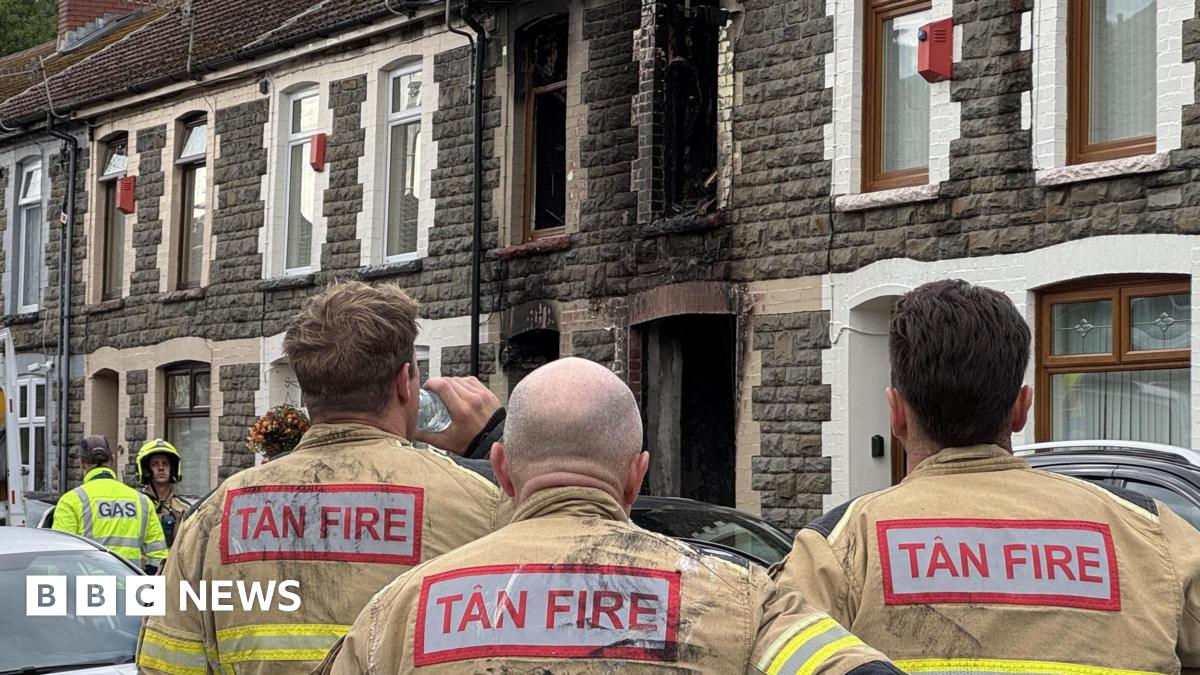 A stone terraced house with fire damage around the windows and doors. Three fire officers look at the house with their backs to the camera.