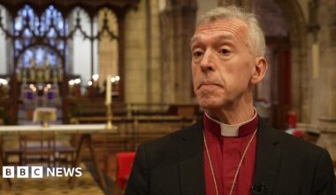 Andrew John in a red shirt and black blazer with a dog collar, looking at the camera in front of an altar. He has grey hair and brown eyes.