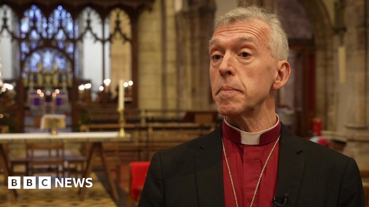 Andrew John in a red shirt and black blazer with a dog collar, looking at the camera in front of an altar. He has grey hair and brown eyes.