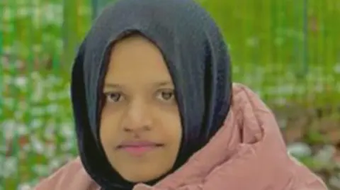 West Yorkshire Police A woman in her 20s wearing a black hijab and pink puffy coat. There is grass behind her although the background is blurred. She has a slightly smiling but fairly neutral expression as she looks at the camera.