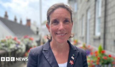 Deputy Lindsay de Sausmarez, a woman with brown hair smiling at the camera. She's wearing a blue jacket and a white blouse. On her lapel are three pin badges.