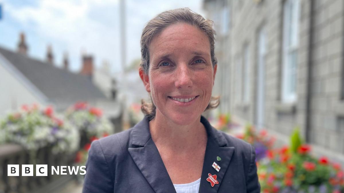 Deputy Lindsay de Sausmarez, a woman with brown hair smiling at the camera. She's wearing a blue jacket and a white blouse. On her lapel are three pin badges.
