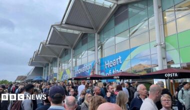 Many people are at the bottom of the photo outside an airport terminal building. Across the image is the building, with a wavy roof  and printed vinyl graphics across the front.