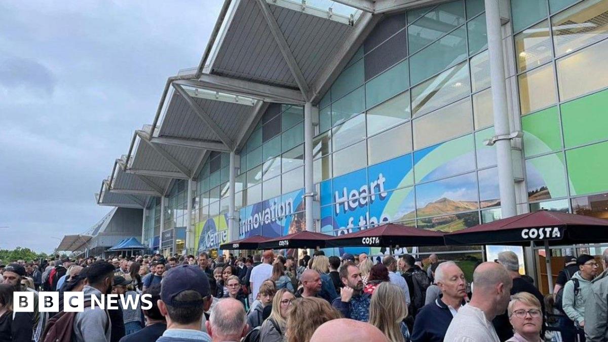 Many people are at the bottom of the photo outside an airport terminal building. Across the image is the building, with a wavy roof  and printed vinyl graphics across the front.