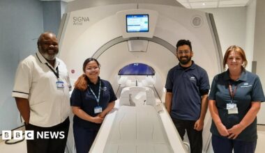 Four members of staff stand around an MRI imaging machine. The trolley where a patient would lie, is outside the actual machine, which has a small circular entrance.