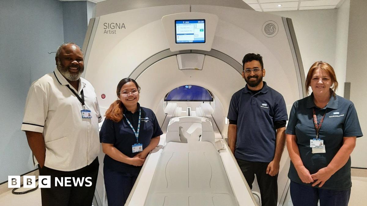 Four members of staff stand around an MRI imaging machine. The trolley where a patient would lie, is outside the actual machine, which has a small circular entrance.