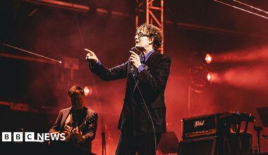 Jarvis Cocker points and sings into a microphone on stage. Next to him is a guitarist.