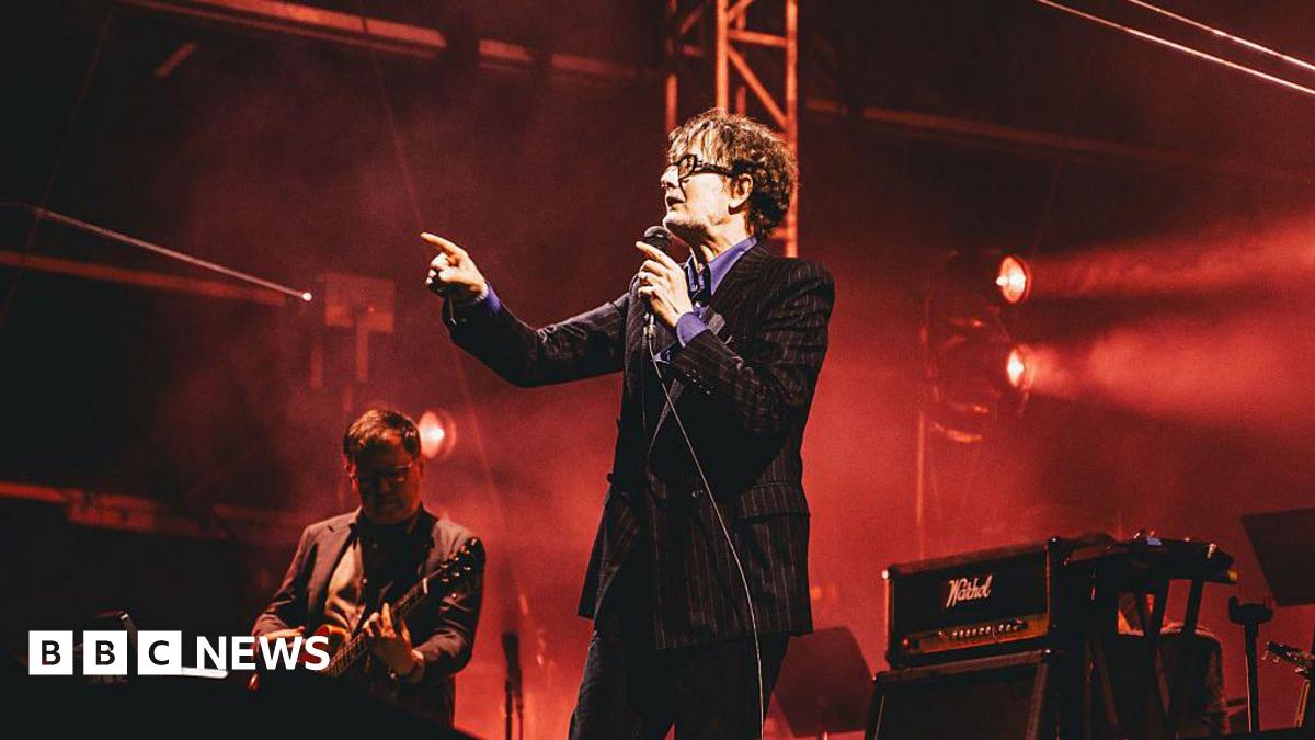 Jarvis Cocker points and sings into a microphone on stage. Next to him is a guitarist.