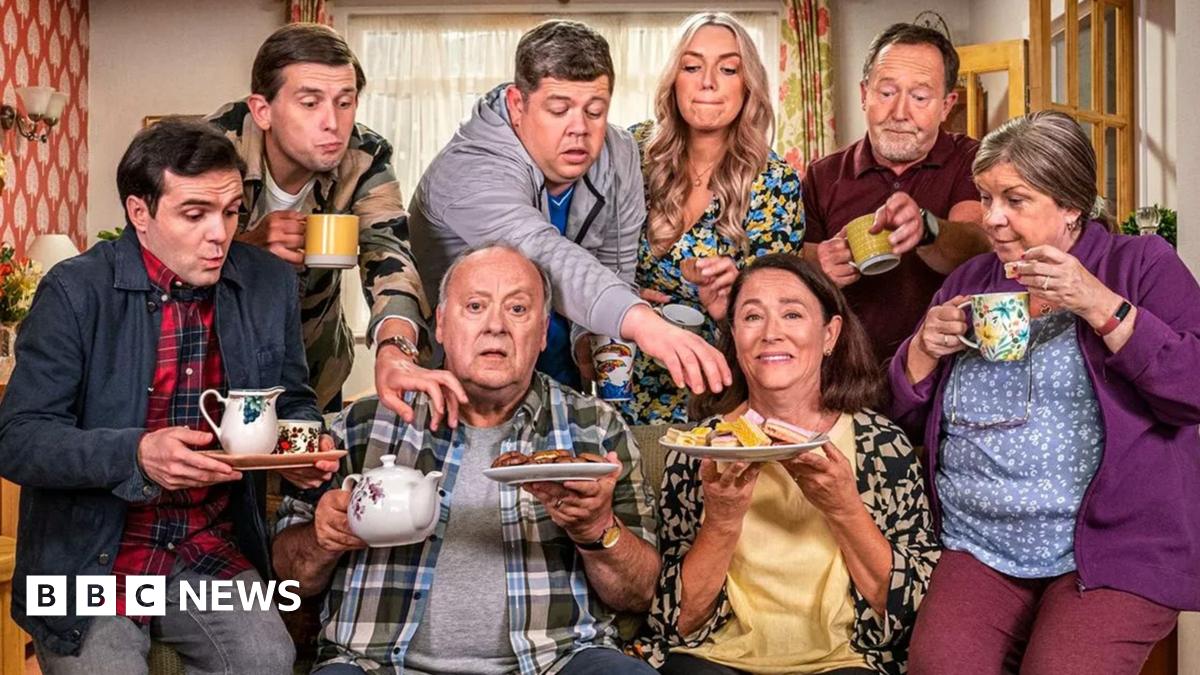 Eight people gathered around a sofa in a living room. Some of them are carrying a selection of mugs, sandwiches and cakes. Two people are on the sofa, two on the arms of the sofa and four are standing behind with two of them reaching out to the plates of food