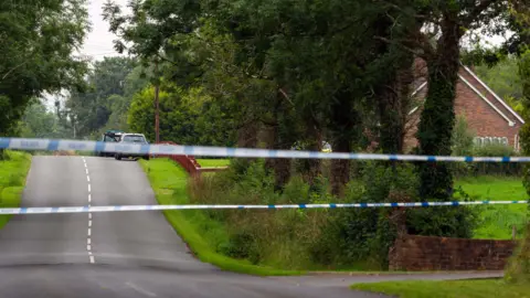 Pacemaker A blue and white police tape across a road with white marking in the middle. Grass verges and trees line the road. In the distance some cars a parked beside a red fence. 