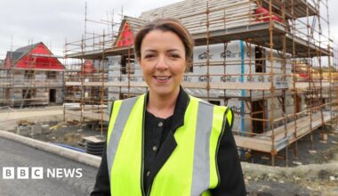 Liz Kimmins stands in front of a house currently under sconstruction. It is covered by scaffolding. Kimmins is wearing a high viz yellow jacket and black blouse. She is smiling and has short brown hair