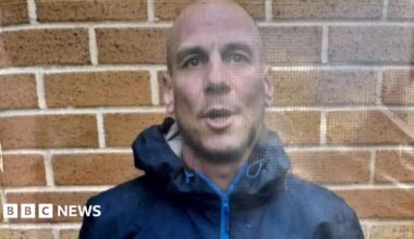 A bald man, who is wearing a dark blue coat with the hood down, has his mouth partially open as he stands in front of a wall.