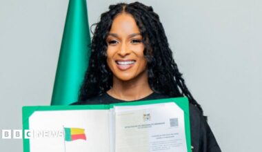 Ciara, wearing a black dress and long black braids, holds up her citizenship documents.