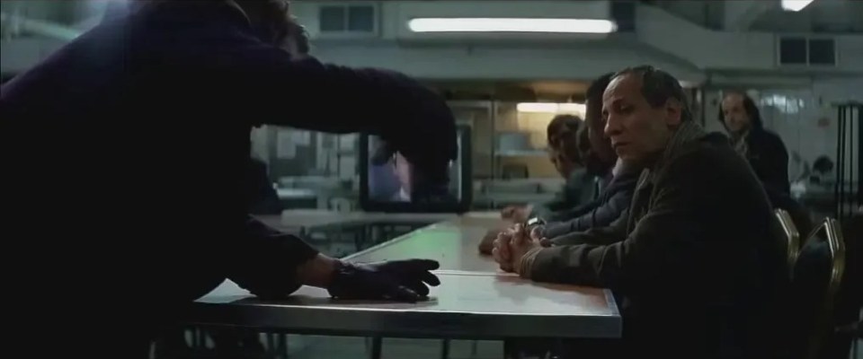 Scene from The Dark Knight: A figure in dark clothing addresses a group of men sitting at a table.