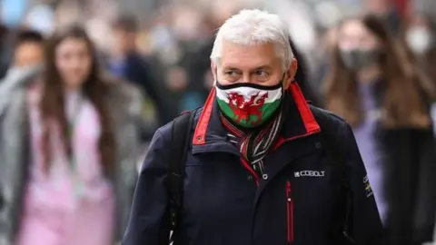 Getty Images Man stood in crowded street wearing a mask with a Welsh flag on it. He is wearing a blue coat and has a rucksack on. People can be seen out of focus behind him.
