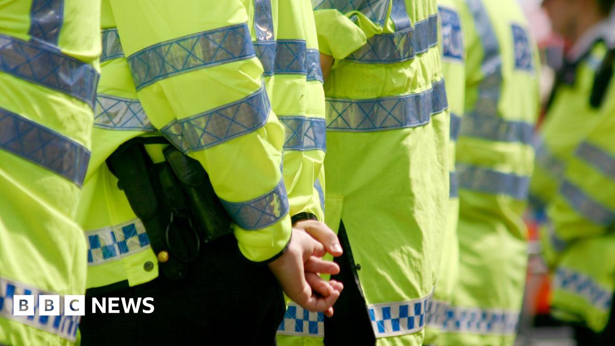 A row of policemen controlling a crowd. Focus is on hands and body of left-most policeman who has his hands interlaced behind his back. The officers are all wearing bright yellow high-vis long-sleeved jackets with silver reflective panelling on them and blue and white check strips along the hem.