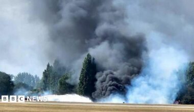 A huge billowing cloud of black, grey and white smoke coming from the edge of the airfield. It is so thick that it is hiding some of the trees - that are on the edge of the airfield. A fire engine can be seen spraying water towards the mass of smoke.
