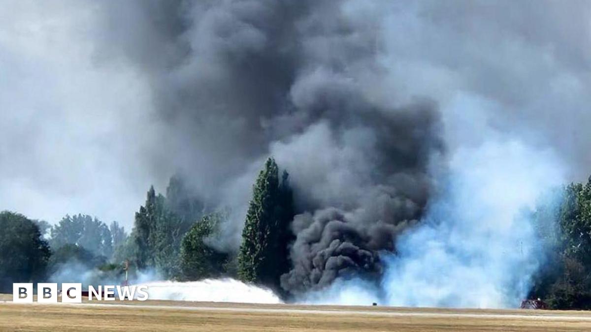 A huge billowing cloud of black, grey and white smoke coming from the edge of the airfield. It is so thick that it is hiding some of the trees - that are on the edge of the airfield. A fire engine can be seen spraying water towards the mass of smoke.