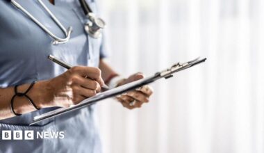 The body of a nurse in blue uniform, holding a clipboard and writing on it. There is a stethoscope around their neck
