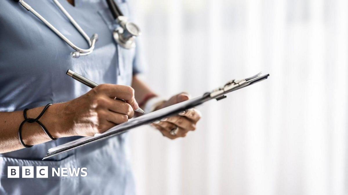 The body of a nurse in blue uniform, holding a clipboard and writing on it. There is a stethoscope around their neck