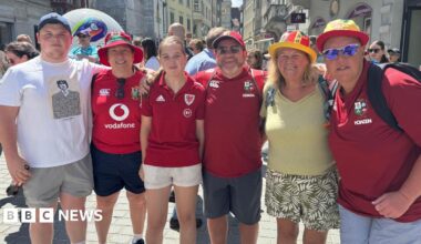 Welsh fans Aneurin Williams, Julie Cashell, Anwen Williams, Stuart Williams, Margaret Rees and Linda Rosser stand in a line with their arms around each other, smiling at the camera. Four of them are wearing red football shirts and Wales bucket hats. They are standing in a street with crowds of people behind them.