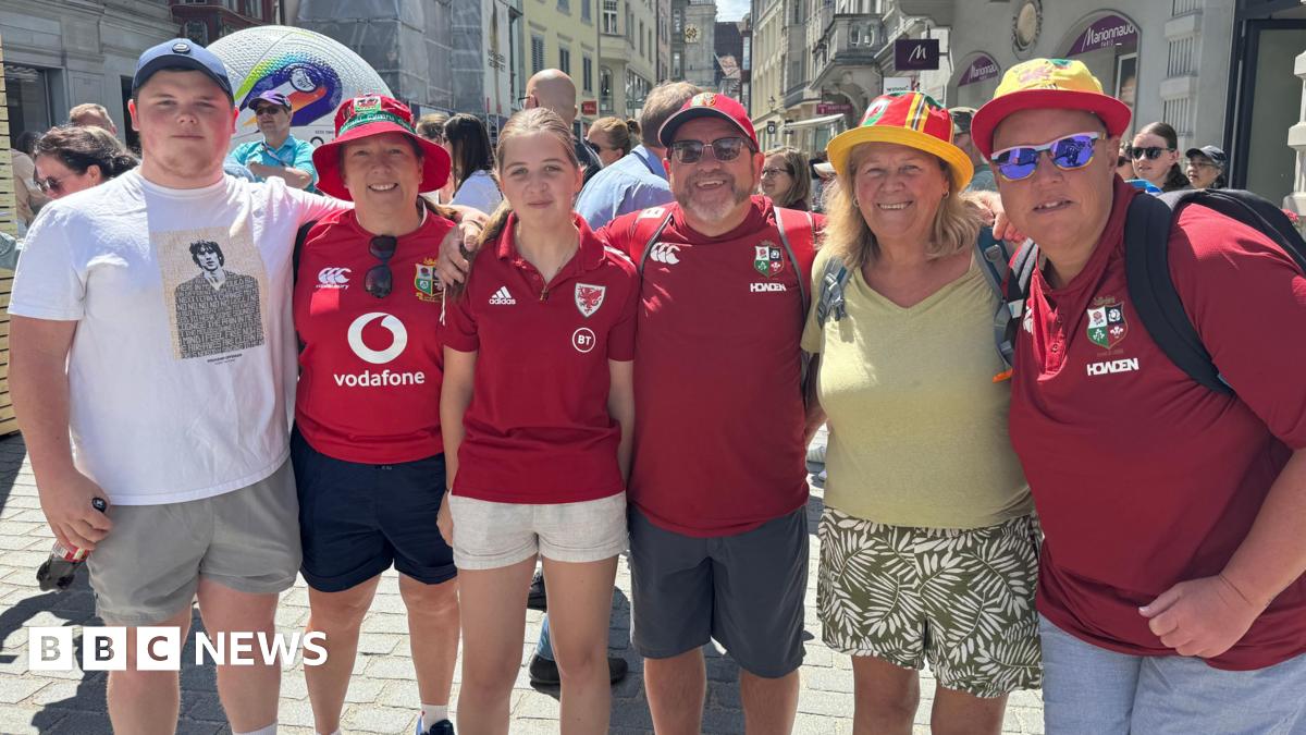 Welsh fans Aneurin Williams, Julie Cashell, Anwen Williams, Stuart Williams, Margaret Rees and Linda Rosser stand in a line with their arms around each other, smiling at the camera. Four of them are wearing red football shirts and Wales bucket hats. They are standing in a street with crowds of people behind them.