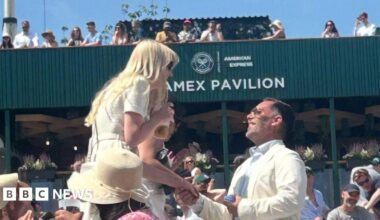 Darren Barsdell on one knee holding Emily Adams' hand and looking up at her as she is surprised looking down while holding a drink. Both are wearing sunglasses and white and cream outfits as they stand in front of one of the pavilions at Wimbledon surrounded by crowds.