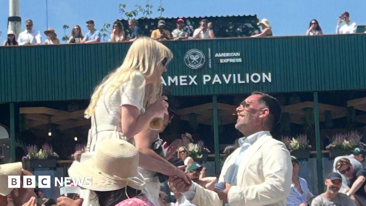 Darren Barsdell on one knee holding Emily Adams' hand and looking up at her as she is surprised looking down while holding a drink. Both are wearing sunglasses and white and cream outfits as they stand in front of one of the pavilions at Wimbledon surrounded by crowds.