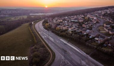Drone footage of Rotherham showing a main road with dozens of houses on one side and fields and greenery on the other.
