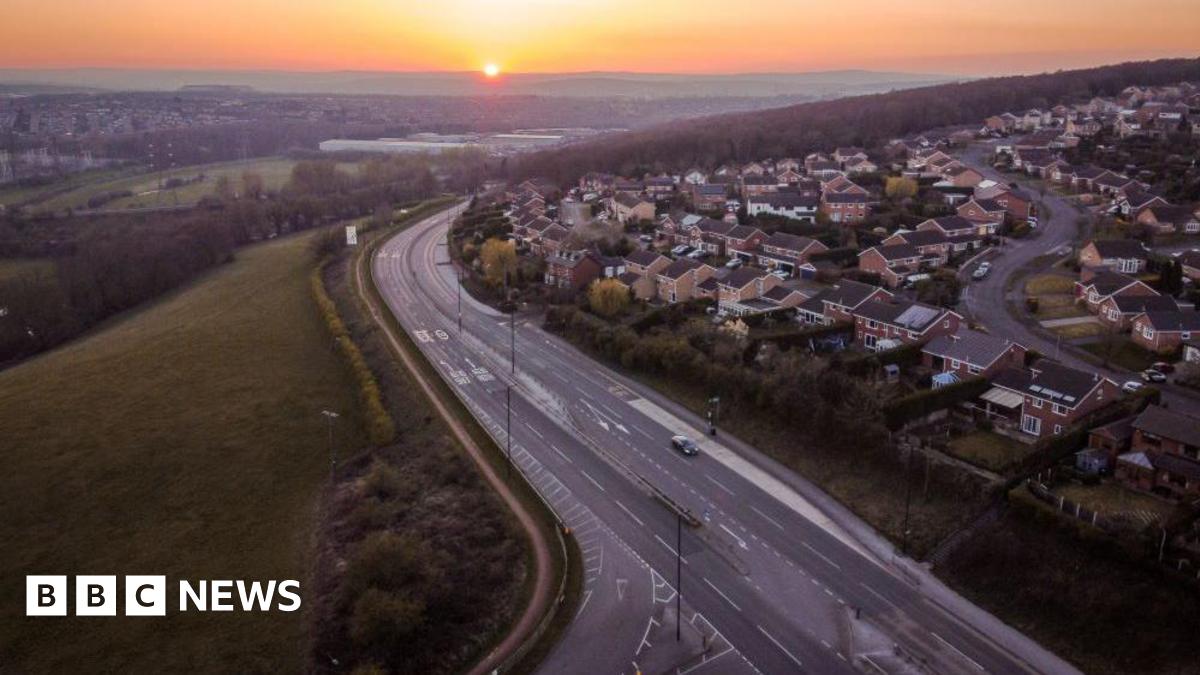 Drone footage of Rotherham showing a main road with dozens of houses on one side and fields and greenery on the other.