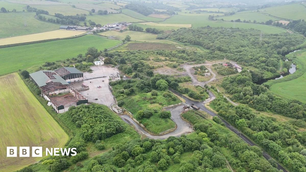 the site of an illegal dump sits surrounded by green fields and countryside. a number of dilapidated buildings are on the left, while minor roads run across the width of the site