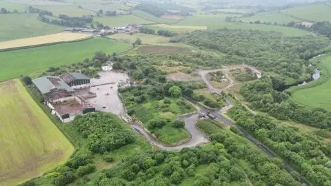 BBC the site of an illegal dump sits surrounded by green fields and countryside. a number of dilapidated buildings are on the left, while minor roads run across the width of the site