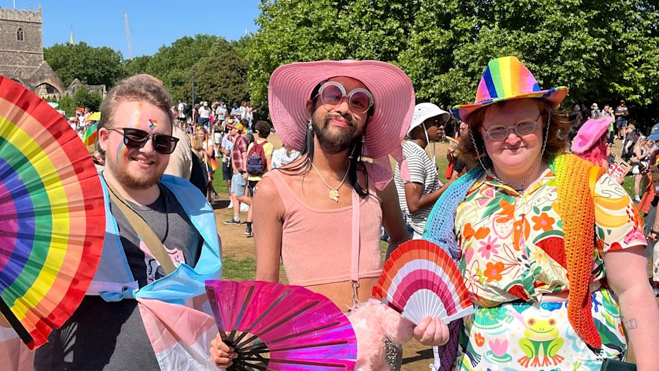 Three attendees smile at the camera. All wearing colourful outfits and holding fans in the heat. Two have facepaint, two have hats, including a rainbow one