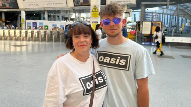 Rachel Rann and her son Thomas are standing side by side. They are both wearing white Oasis T-shirts and are smiling at the camera.