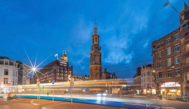 A cityscape at dusk shows blurred lights from a passing tram in front of historic buildings and a tall clock tower, with a deep blue sky in the background.